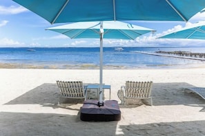 Un entorno de playa con dos sillas de mimbre bajo un paraguas grande, una mesa y una guitarra.