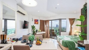 Una habitación de hotel moderna con zona de comedor, cama con edredón verde y un amplio ventanal con vistas a la ciudad.