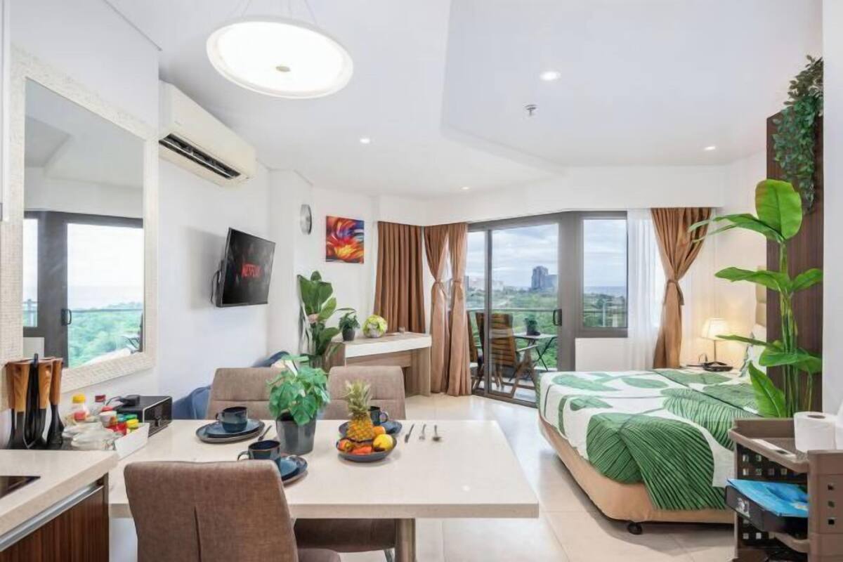 Una habitación de hotel moderna con zona de comedor, cama con edredón verde y un amplio ventanal con vistas a la ciudad.