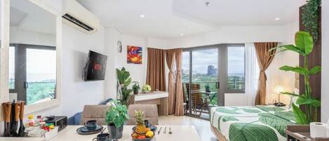 Una habitación de hotel moderna con zona de comedor, cama con edredón verde y un amplio ventanal con vistas a la ciudad.