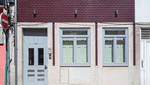 En bygning med en grå dør markeret med nummer 26, to vinduer med lysegrønne skodder, og en hvid dør til højre.