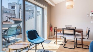 A modern living room with a large sliding glass door leading to a balcony, a dining area with a wooden table and chairs, and a blue chair.