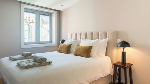 A neatly made bed with white and brown pillows, a bedside lamp, and a window with a view of buildings.