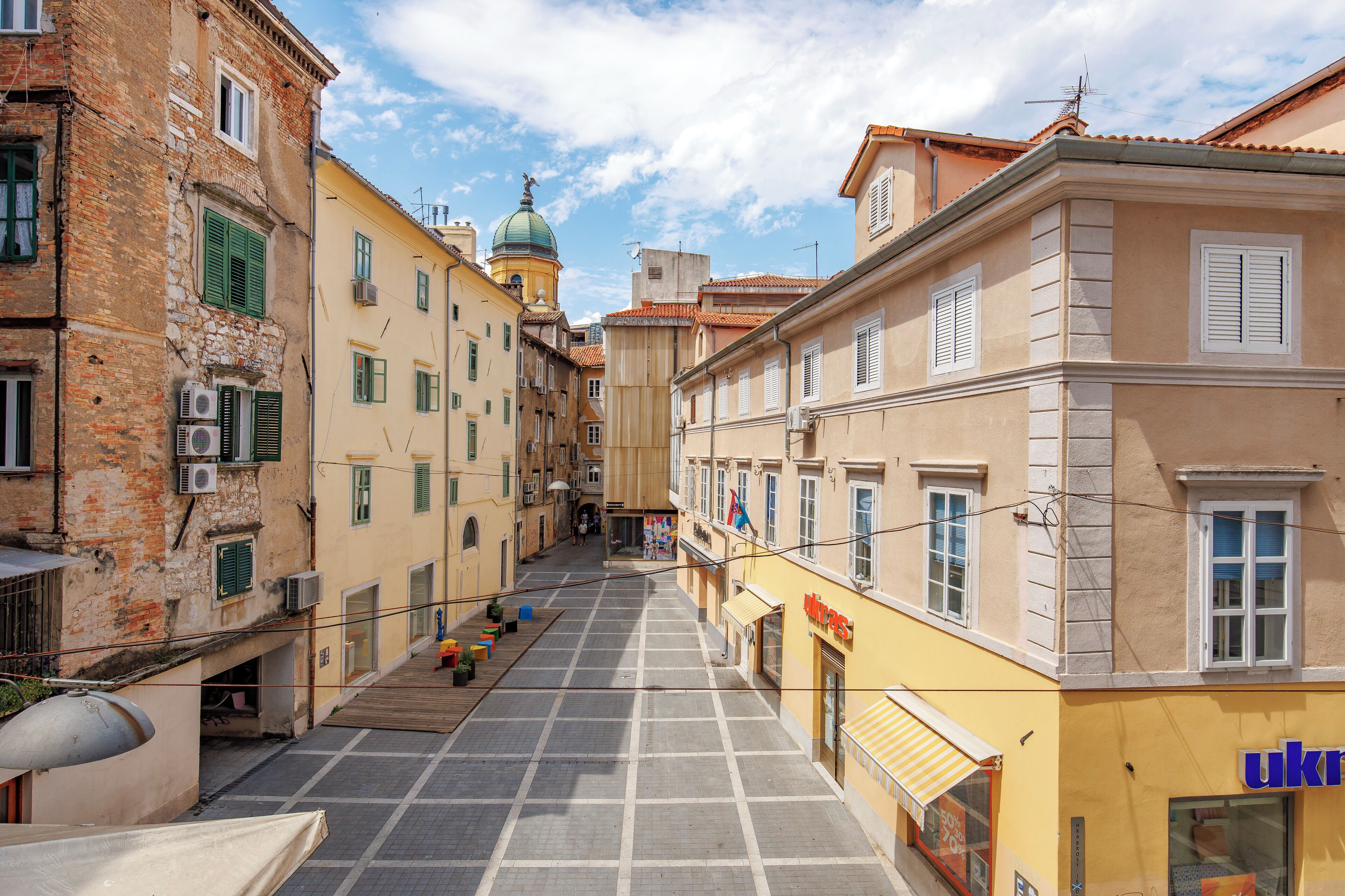 A narrow street with buildings on either side, a clear sky, and a few people walking.