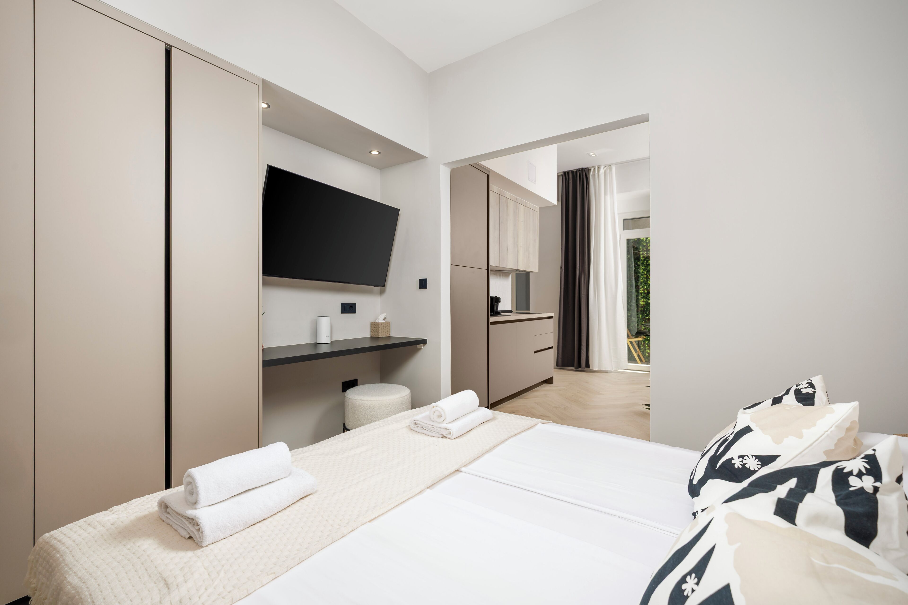 Una habitación de hotel moderna con una cama, un televisor montado en la pared, una pequeña cocina y vistas al exterior.