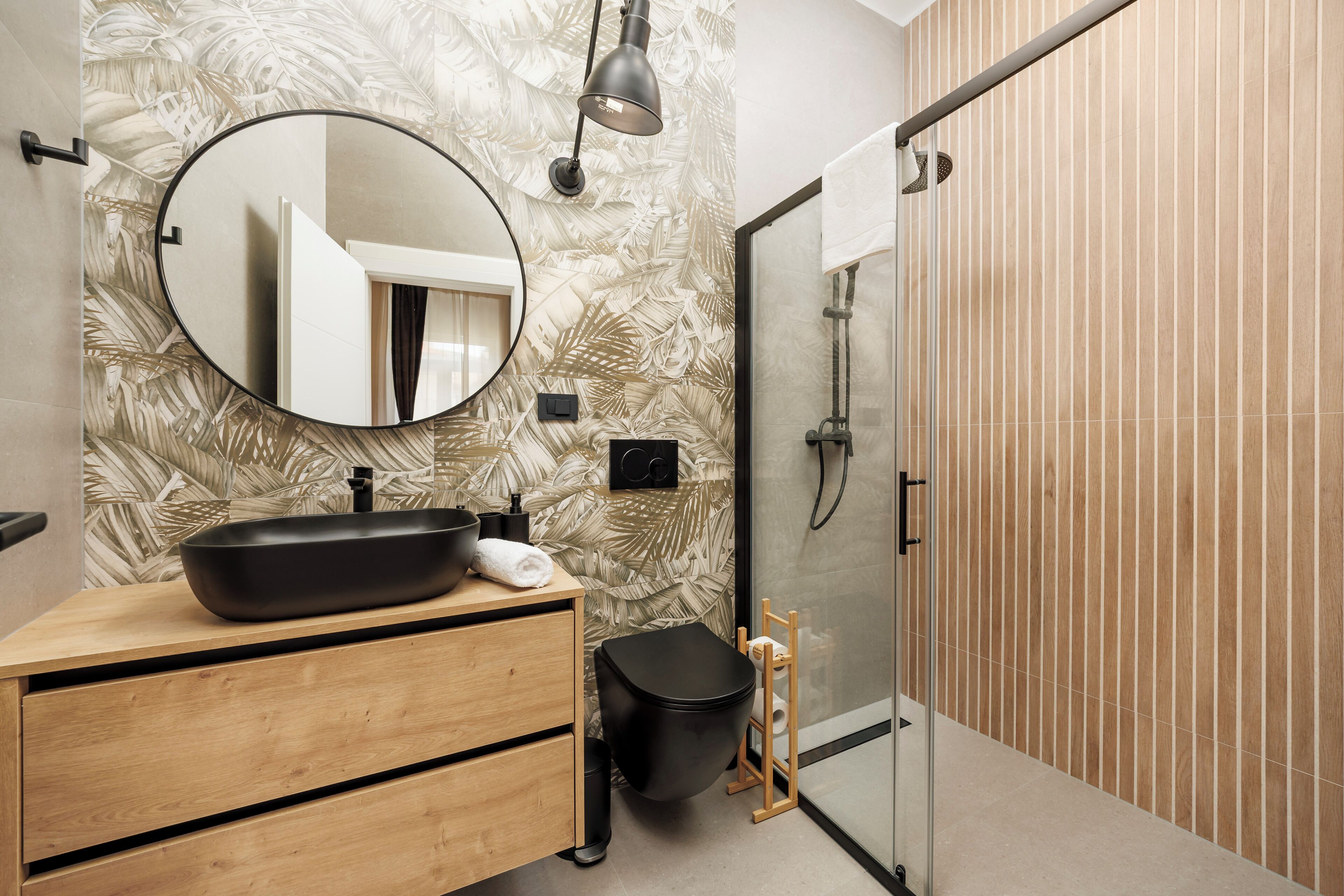 A modern bathroom with a round mirror, a black sink, wooden drawers, a black toilet, and a glass shower enclosure.