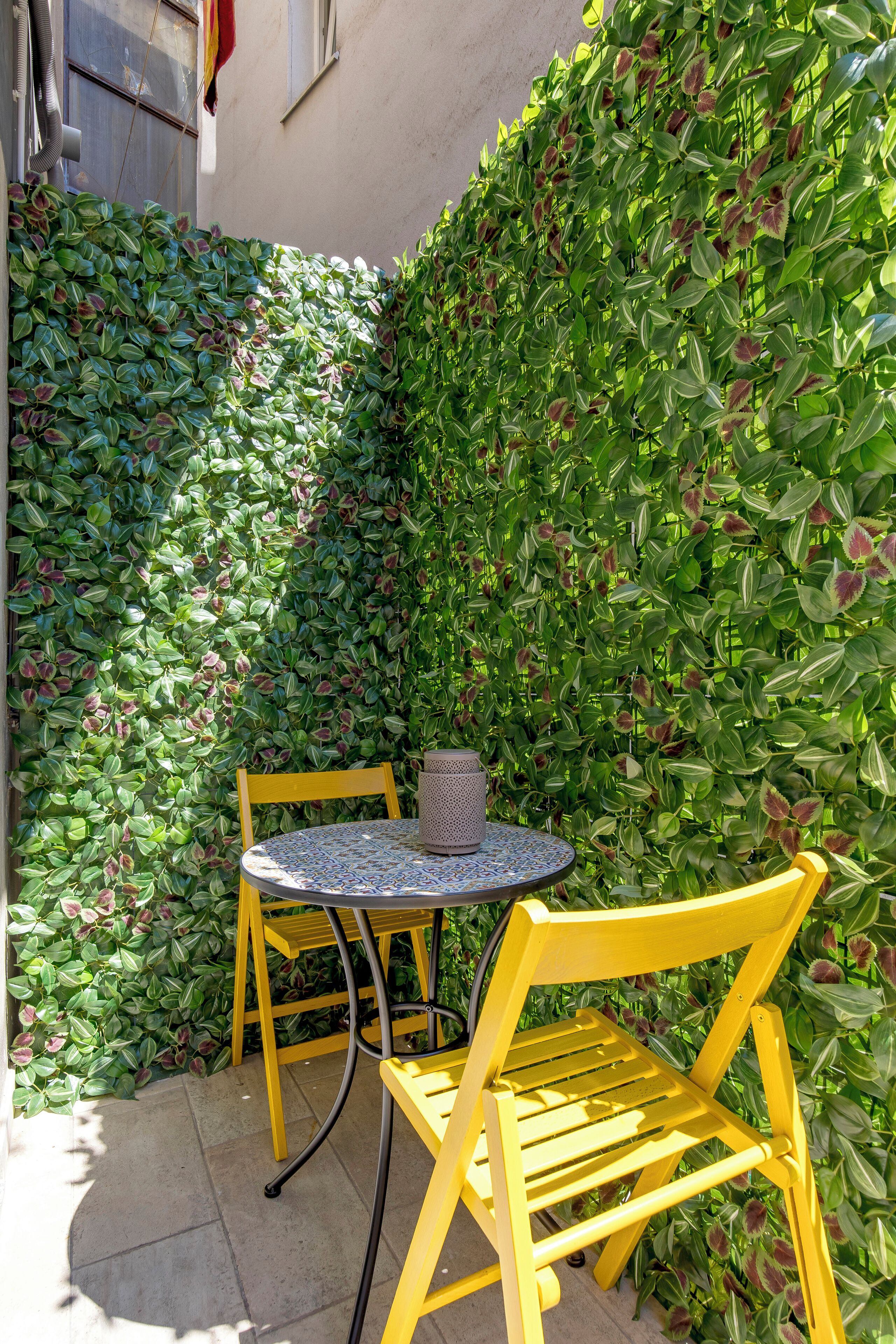 A small outdoor seating area with a round table and two yellow chairs, surrounded by a dense green hedge.
