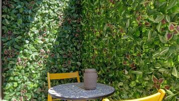 A small outdoor seating area with a round table and two yellow chairs, surrounded by a dense green hedge.