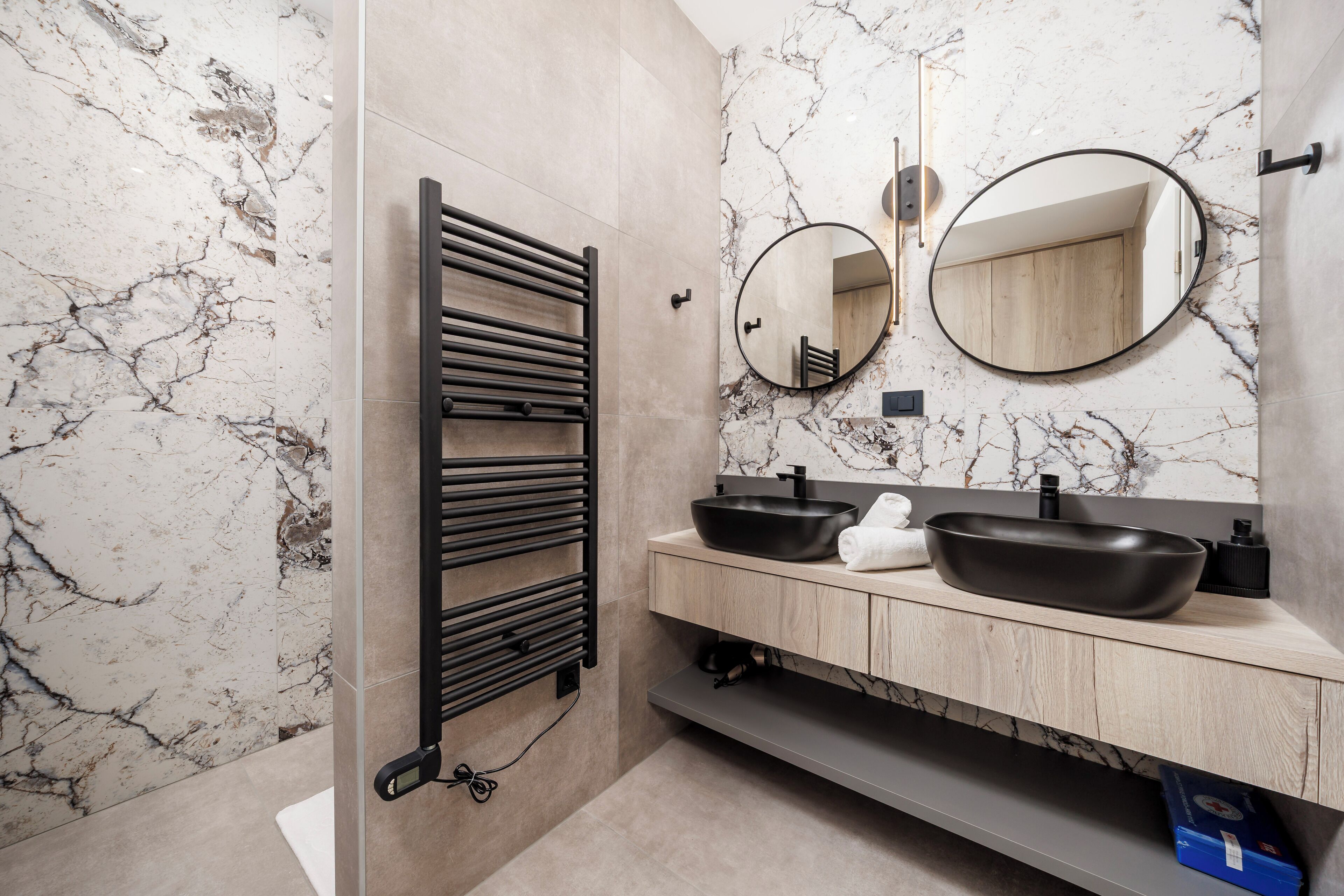 A modern bathroom with marble walls, a black towel rack, two sinks, and a mirror.
