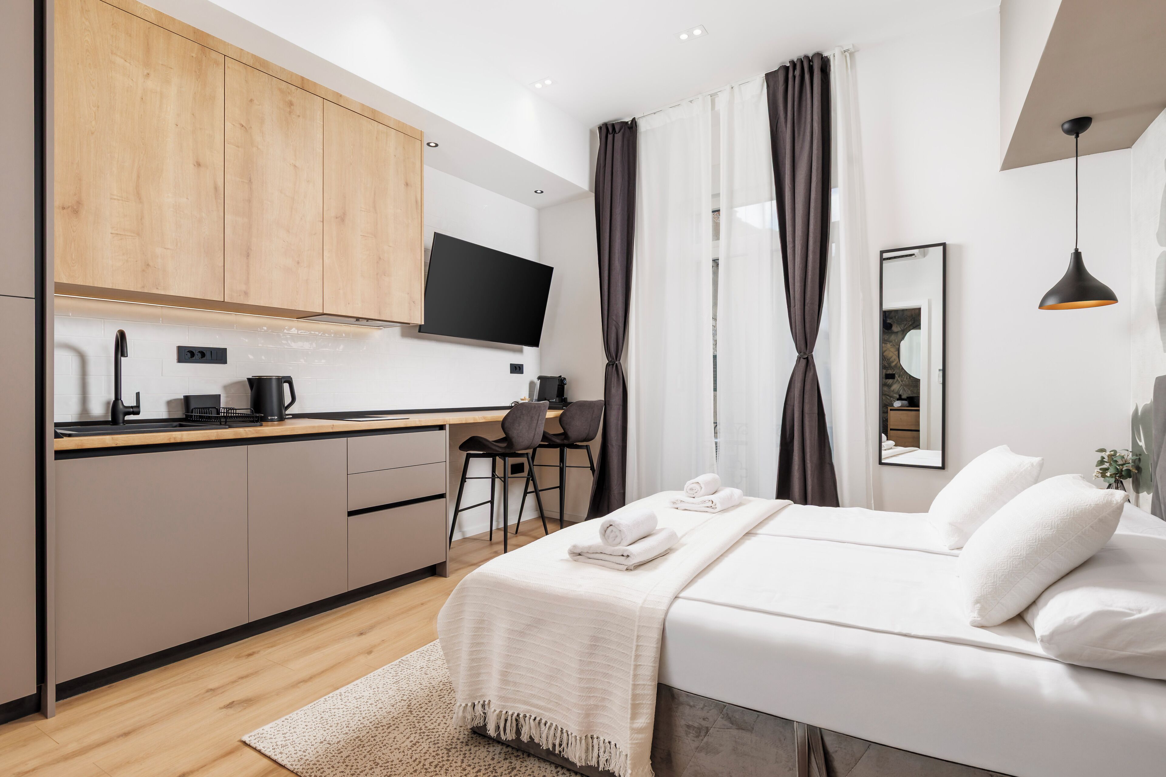 A modern hotel room with a wooden kitchen, a flat-screen TV, a bed with white linens, and a large mirror.