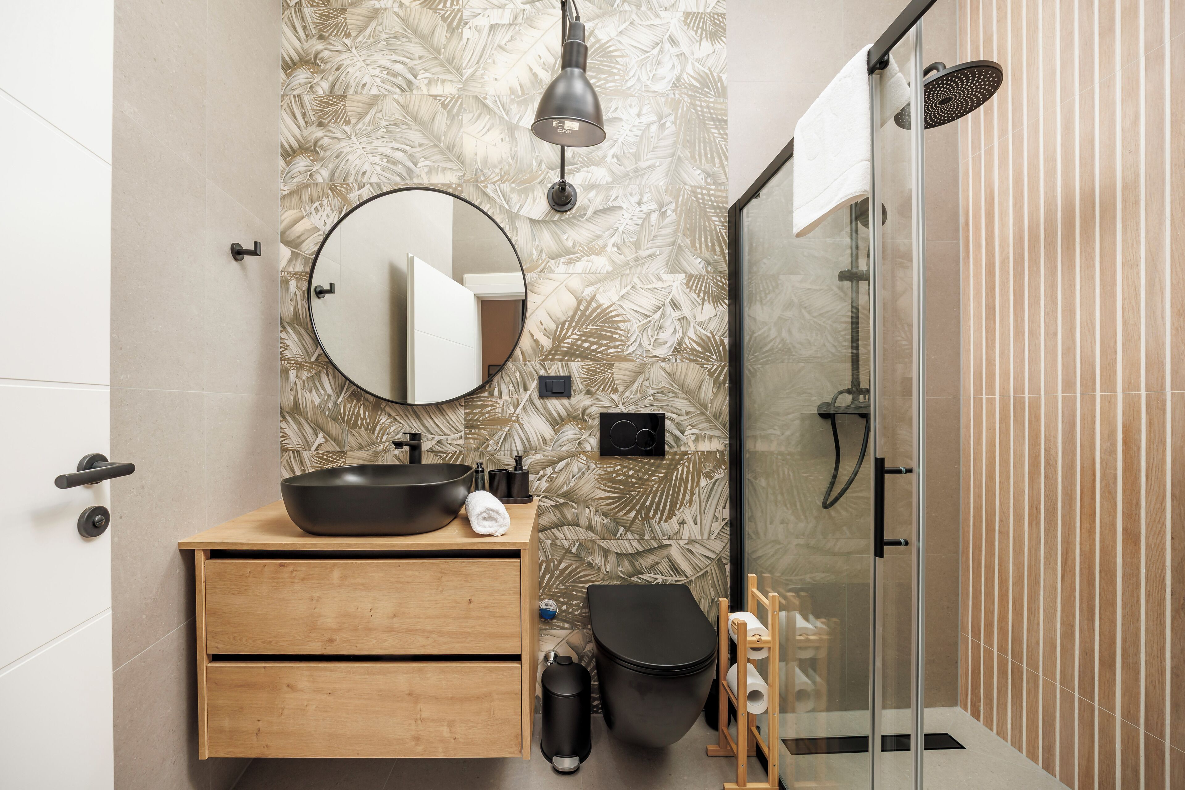 Une salle de bain moderne avec un meuble-lavabo en bois, un miroir rond, un lavabo noir, des toilettes et une cabine de douche en verre.