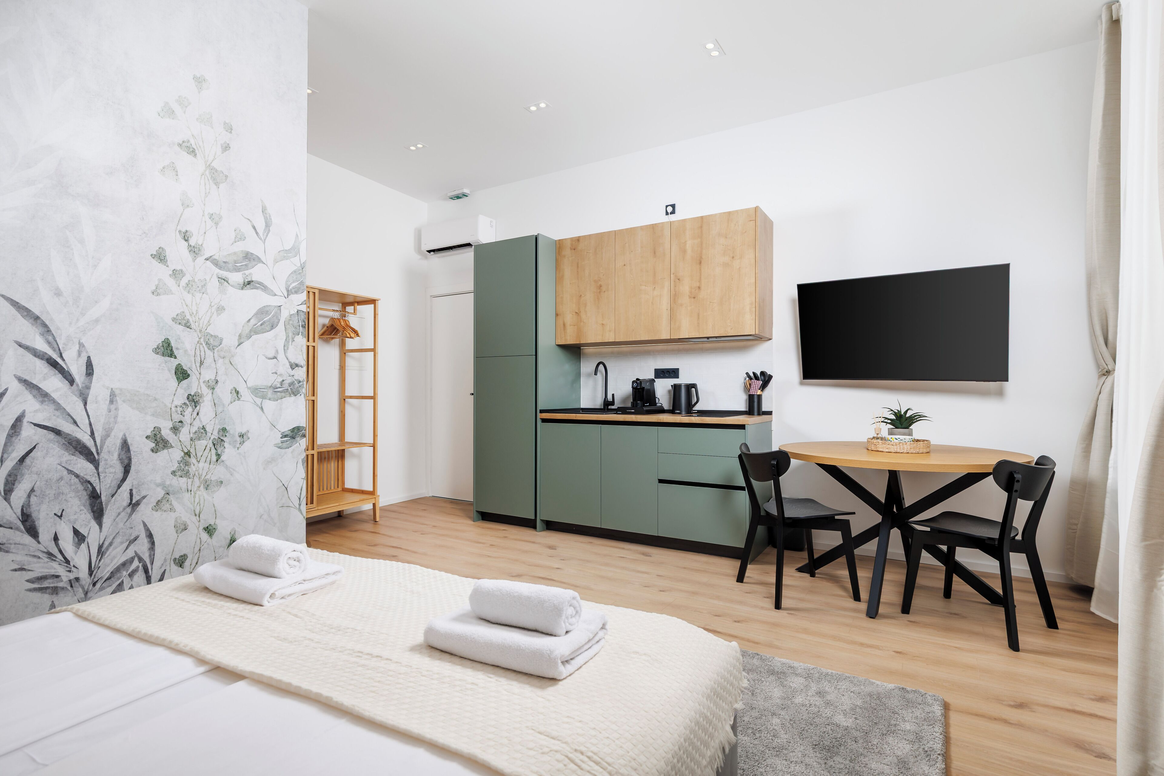 A modern hotel room with a bed, a small kitchenette, a dining area, and a TV.