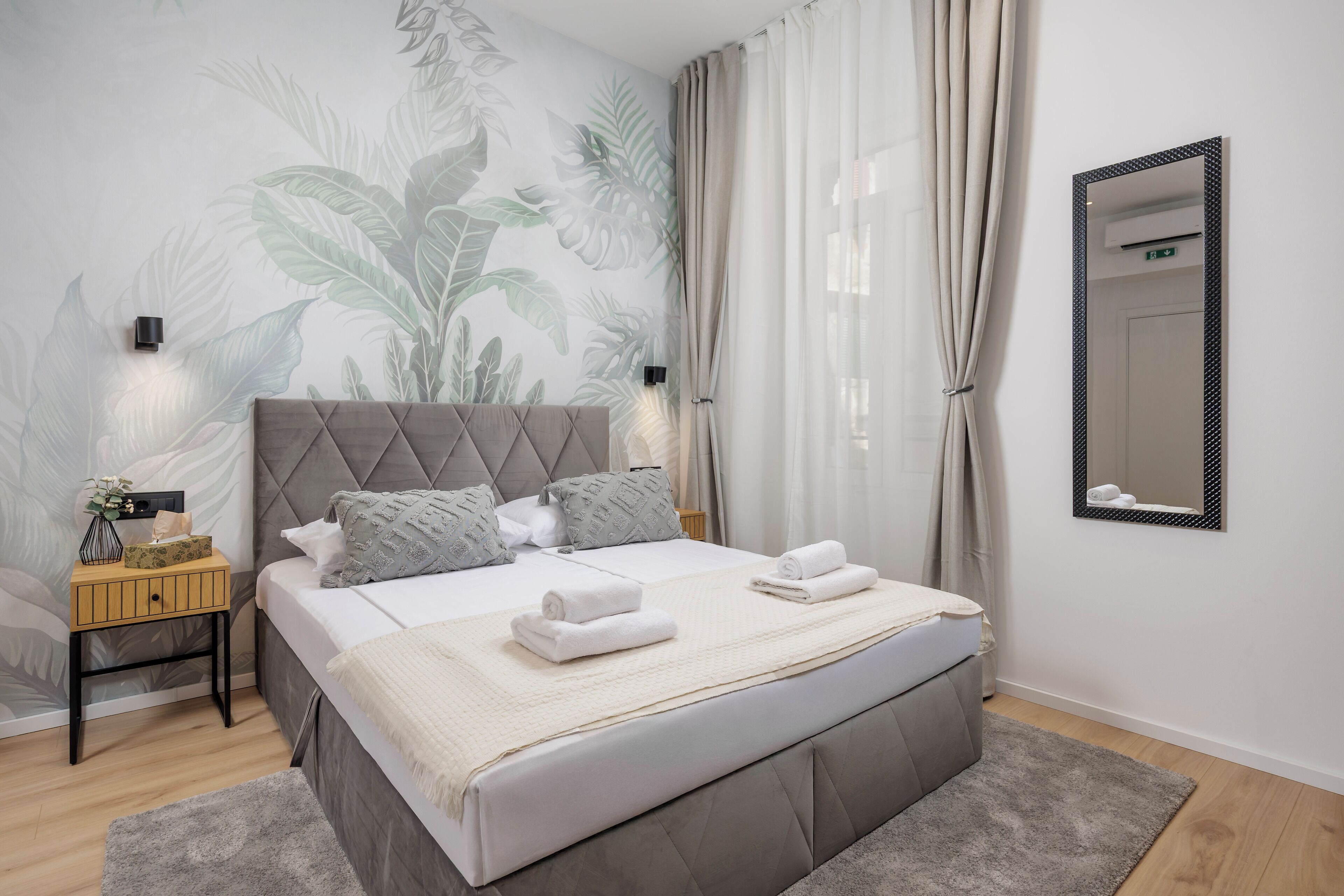 A modern bedroom with a large bed, a nightstand, a wall-mounted mirror, and a tropical-themed wall mural.