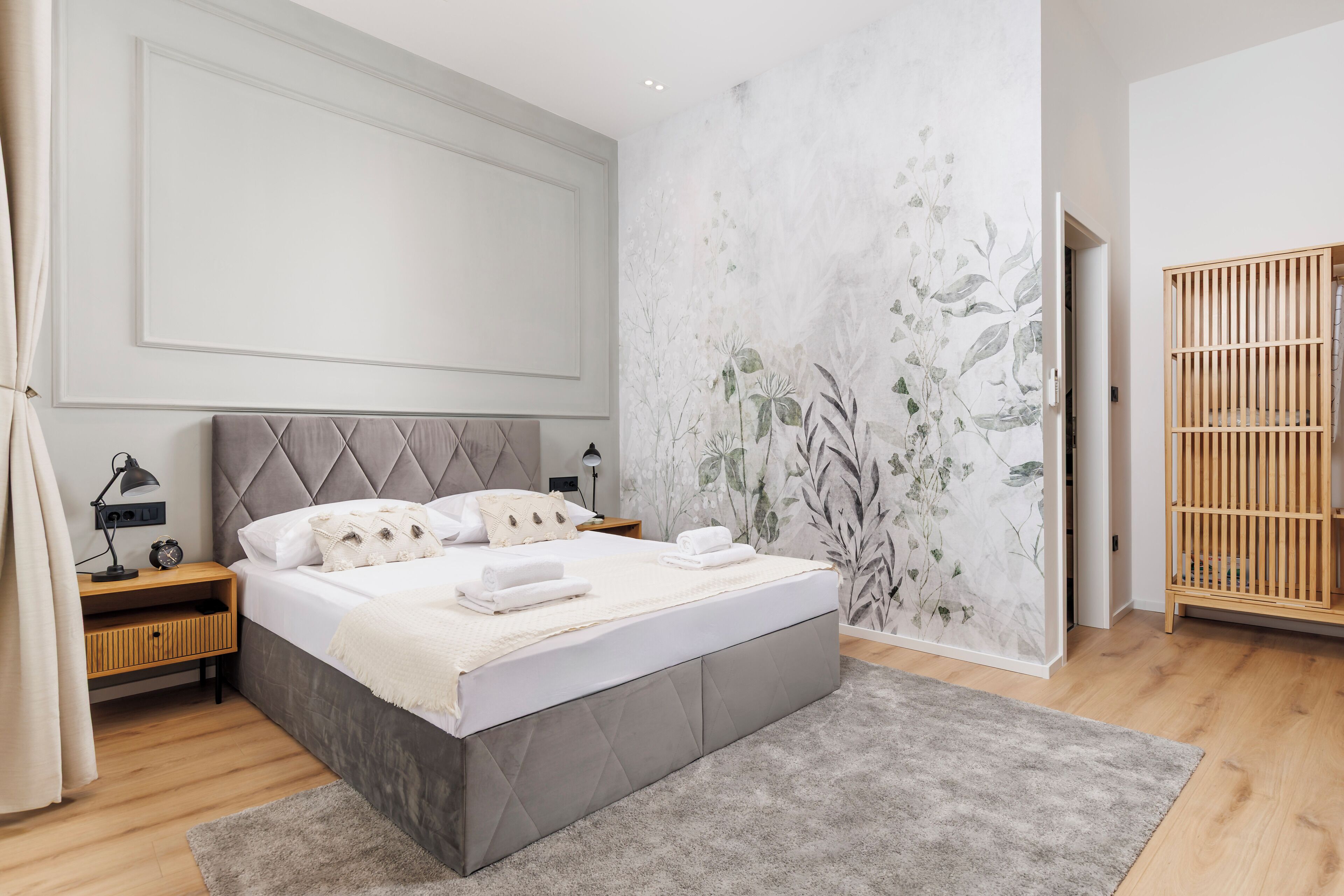 A modern bedroom with a large bed, wooden nightstands, a grey upholstered headboard, and a floral wallpaper design.