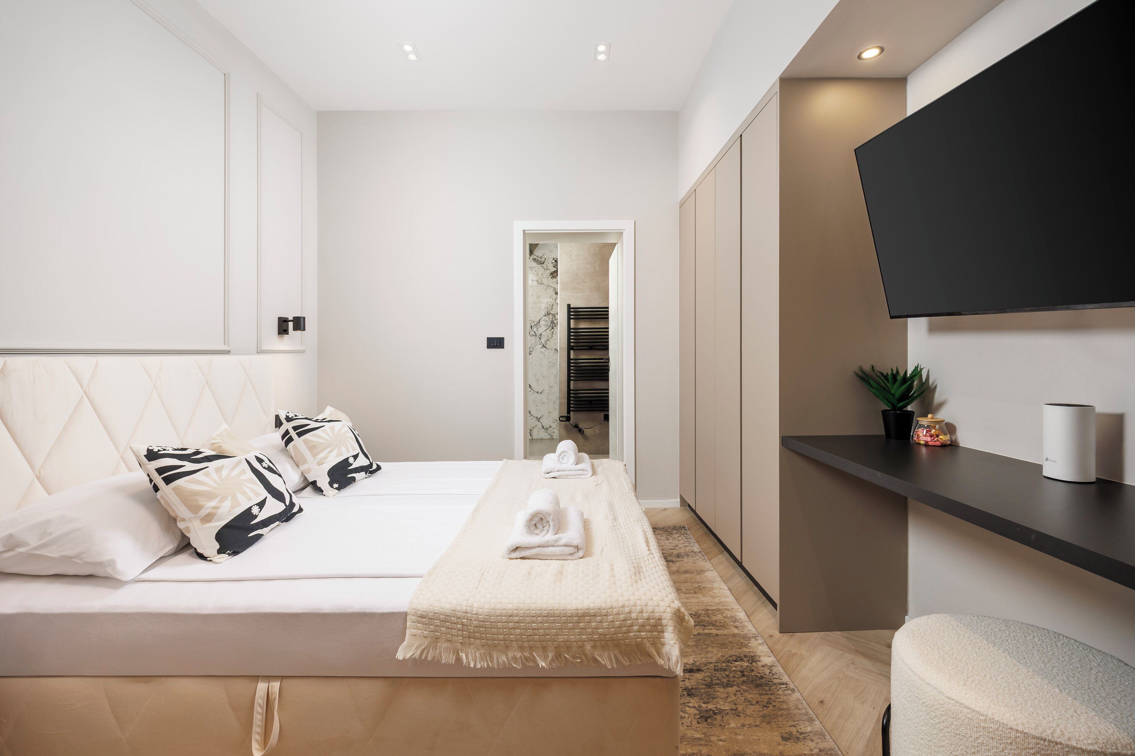 A modern bedroom with a large bed, a flat-screen TV mounted on the wall, and a small plant on a shelf.