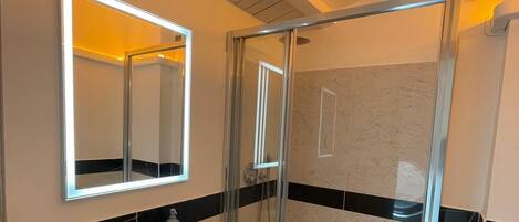 Ein modernes Badezimmer mit einer Duschkabine, einem weißen Waschbecken und einem großen, beleuchteten Spiegel.