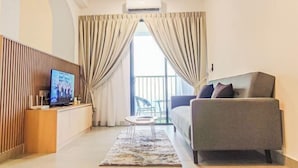 A modern hotel room with a sofa, a TV on a wooden console, a small table, and a view through the curtains to an outdoor area. - Mall Suite at Sunsuria Setia Alam by HCK (Petaling)