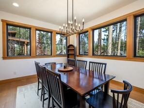 Un coin repas avec une table et des chaises en bois, de grandes fenêtres donnant sur la verdure et un lustre.