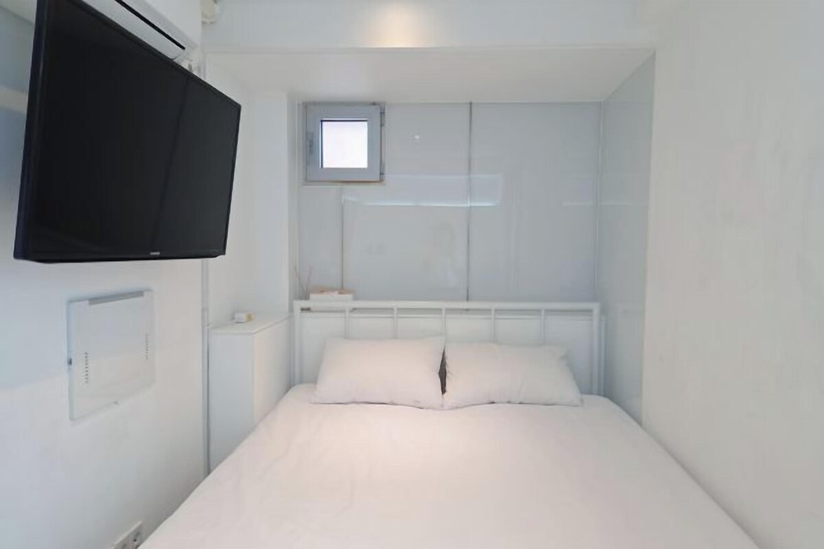 A small, white, single-bed room with a mounted TV, a small window, and a white wardrobe.