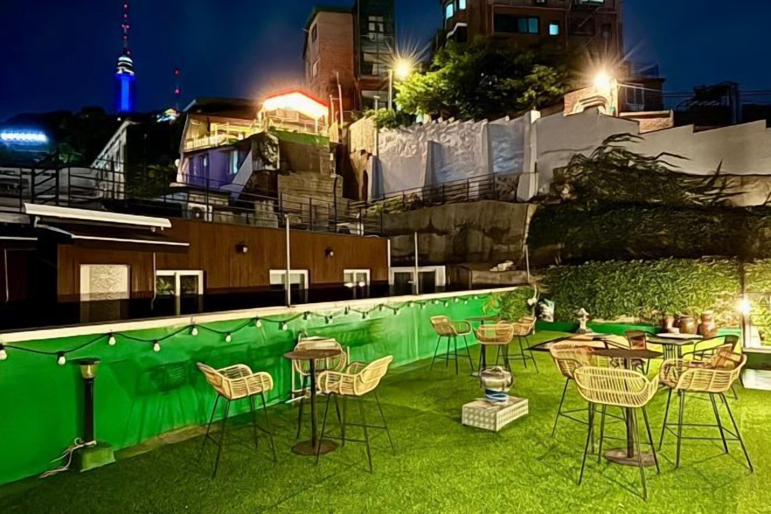 An outdoor seating area with tables and chairs, surrounded by a green hedge and a building with a staircase.