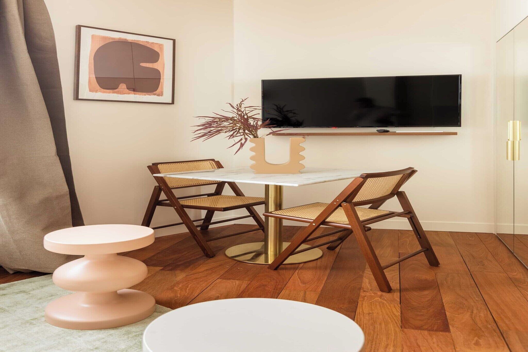 A modern living room with a round table, two wooden chairs, a small stool, a TV mounted on the wall, and a framed artwork.