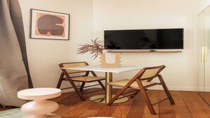 A modern living room with a round table, two wooden chairs, a small stool, a TV mounted on the wall, and a framed artwork.