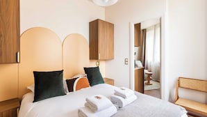 A neatly made bed with white linens, two green pillows, and a grey throw. There are two wooden cabinets above the bed, a wooden bench to the right, and a mirror on the wall.