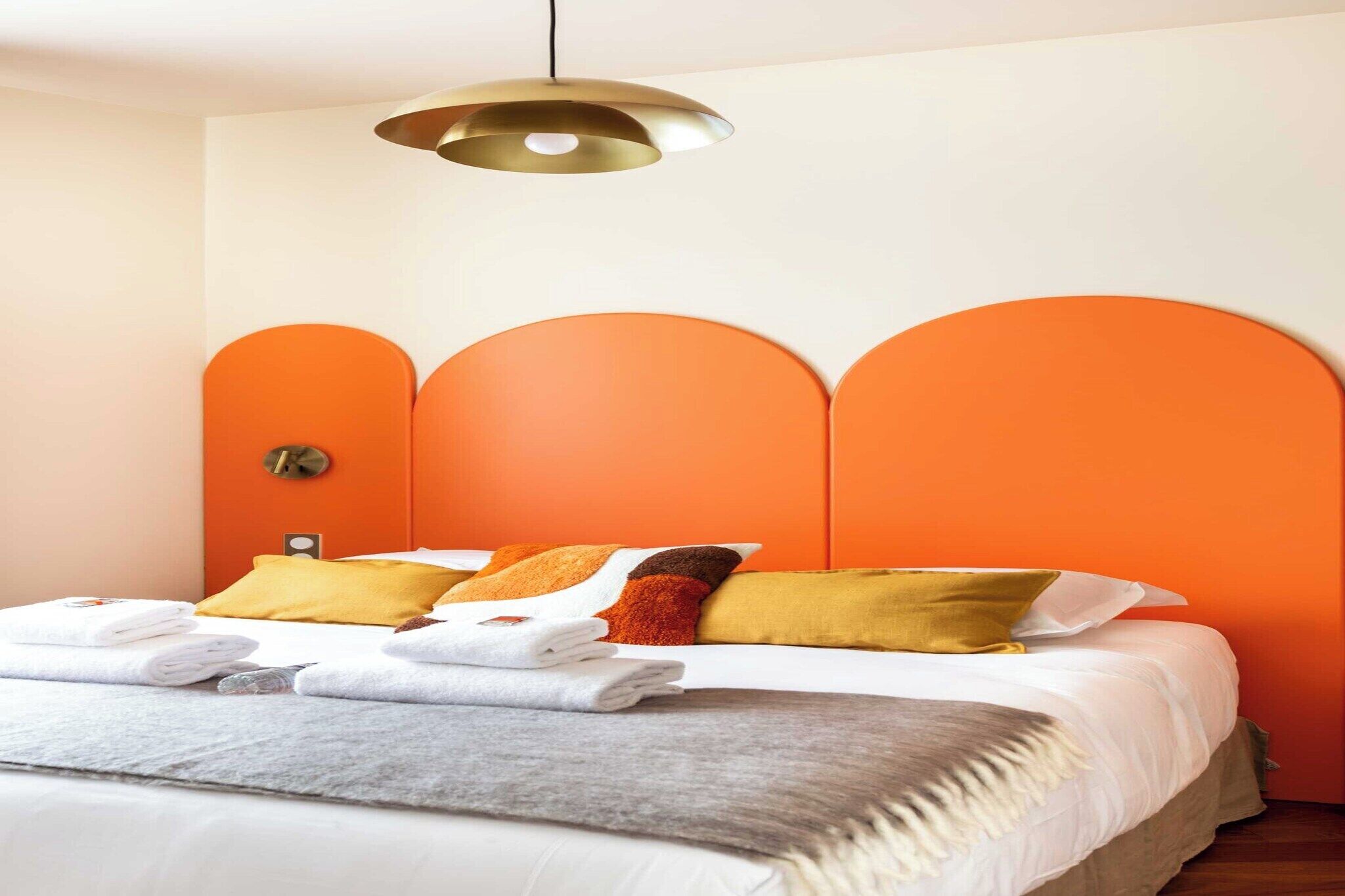 Ein ordentlich bezogenes Bett mit weißen Bettwäsche und einer grauen Tagesdecke, orangefarbenen Kissen und einer goldenen Pendelleuchte darüber.