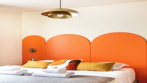 A neatly made bed with white linens and a grey throw blanket, orange accent pillows, and a gold pendant light above. - Port-Royal Residence - Chic apartment - 2BR-4P (Paris)