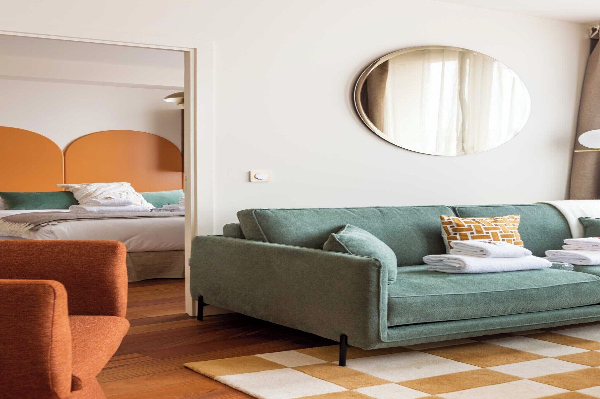 A modern hotel room with a green sofa, a wooden floor, a checkered rug, and a bed with a white headboard.
