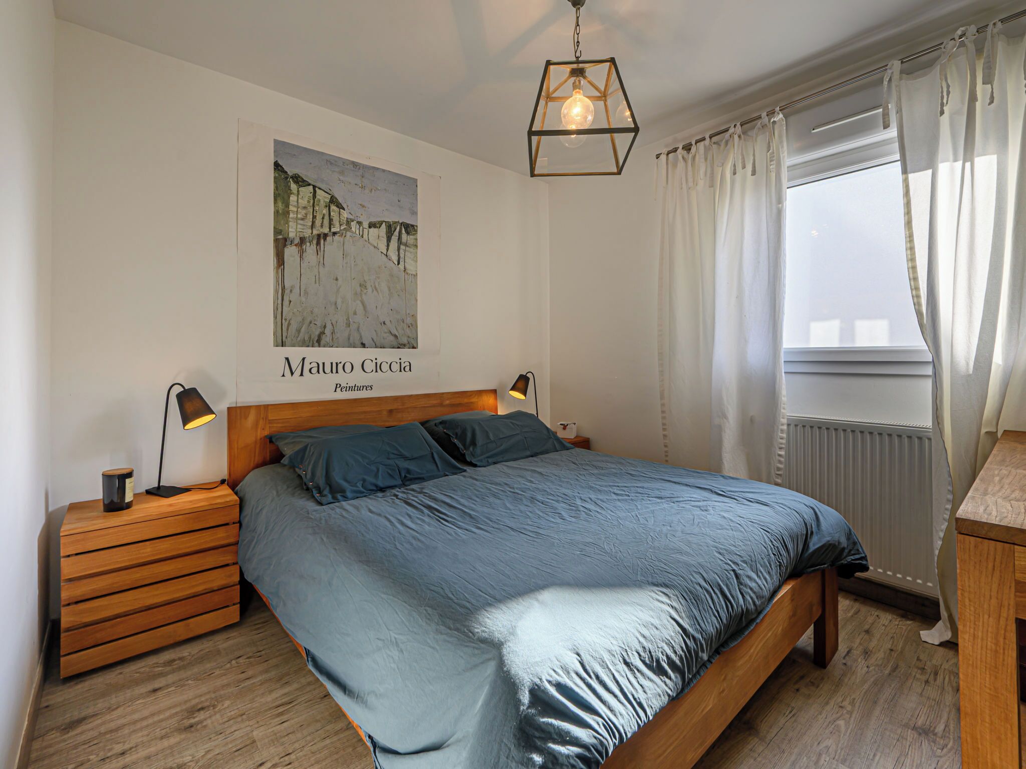 Een slaapkamer met een houten bed, twee bedlampen, een nachtkastje, een raam met gordijnen en een schilderij van een besneeerd landschap.