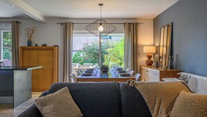 A modern living room with a dark grey sofa, a dining area with a table set for four, and a view of a garden through large windows. - Maison Er Menhir by Interhome (Plouhinec Morbihan)