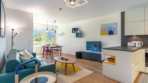 Un apartamento moderno con sala de estar, comedor y cocina. Hay un sofá, una mesa de centro y un televisor. La cocina tiene gabinetes blancos y una encimera.
