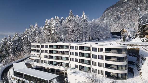 Un hotel moderno de varios pisos con techo plano, rodeado de árboles cubiertos de nieve y una montaña al fondo.