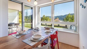 Un comedor con una mesa de madera dispuesta para dos personas, ventanas amplias con vistas a un paisaje verde y lámparas colgantes.