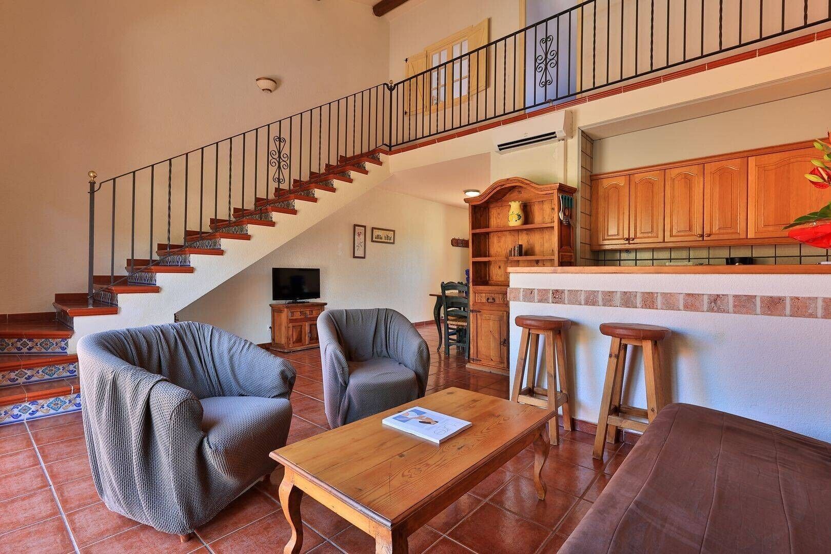 Un salon avec un escalier en bois, deux fauteuils, une table basse et un coin cuisine doté de meubles en bois.