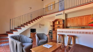 A living room with a wooden staircase, two armchairs, a coffee table, and a kitchen area with wooden cabinets. - Villa '23 Maria Callas' with Private Terrace, Wi-Fi and Air Conditioning (Ramatuelle)