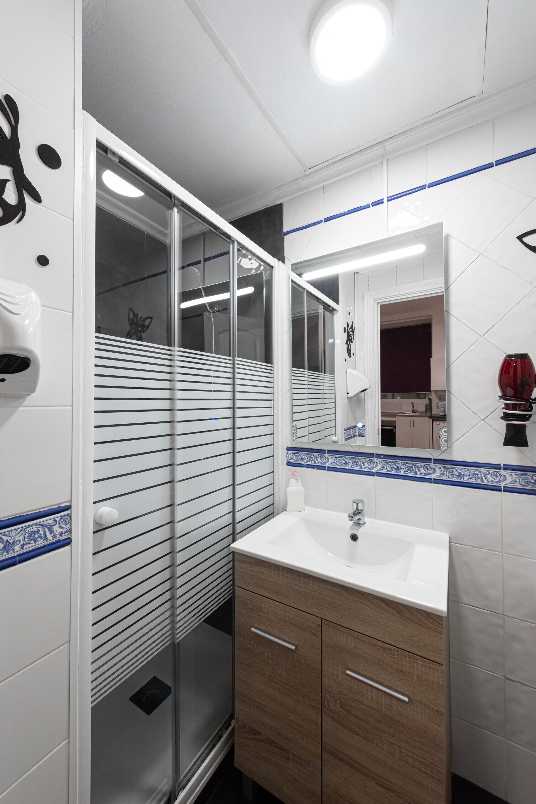 Un baño moderno con cabina de ducha de vidrio, un lavamanos blanco con mueble de madera y un espejo que refleja la habitación.