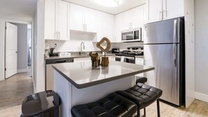 Una cocina moderna con armarios blancos, un refrigerador de acero inoxidable, un microondas y una máquina de café. Cuenta con una barra con dos taburetes.