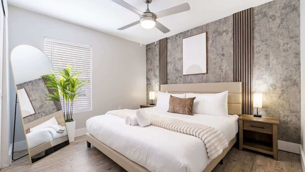 Un dormitorio moderno con una cama grande, un ventilador de techo, una mesita de noche con lámpara y un espejo amplio.