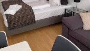A single bed with a white mattress and a brown throw, a nightstand with a lamp, a purple sofa, and a small table with chairs. - Marsktures motel (Højer)