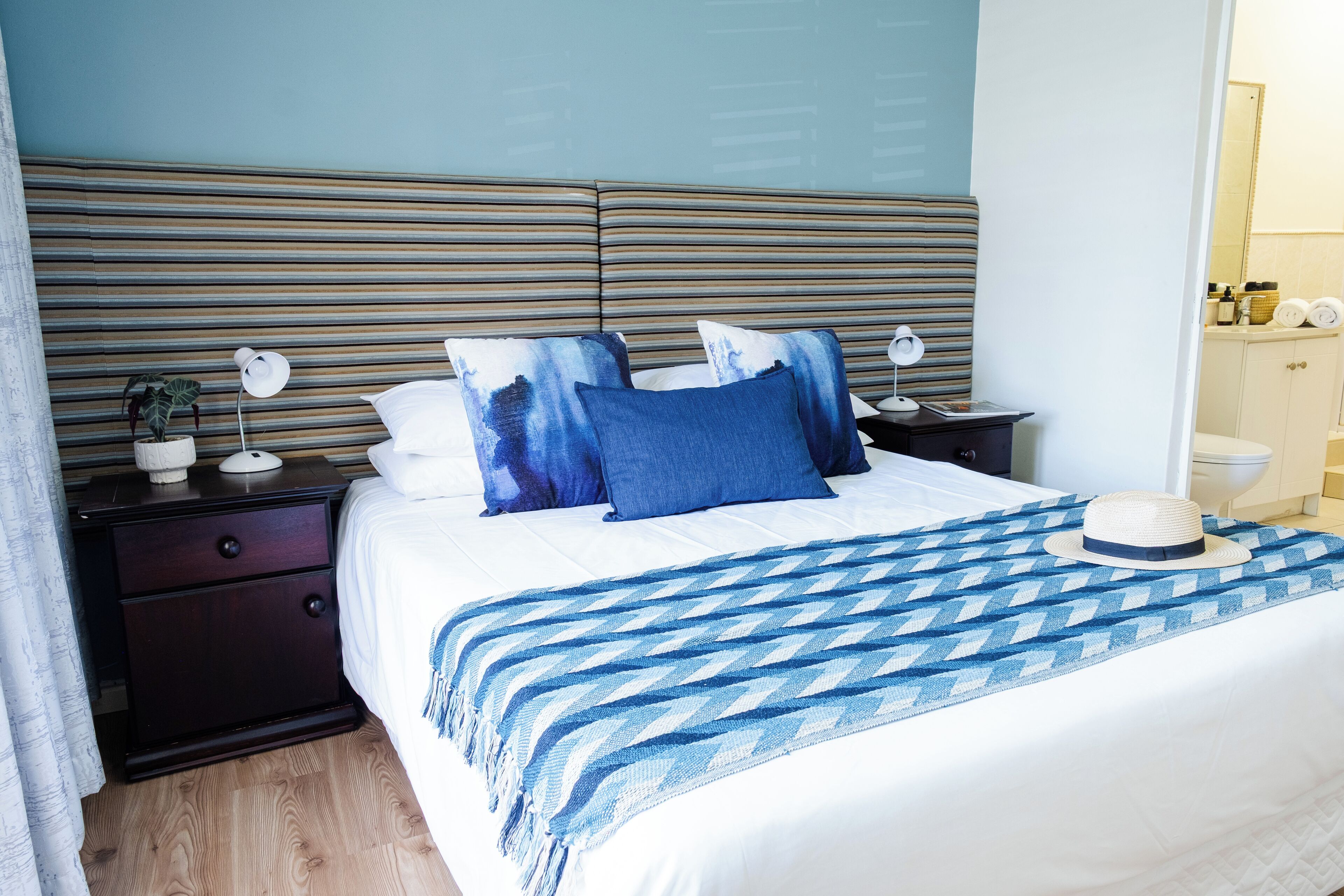 Una cama bien hecha con cabecera a rayas, una mesita de noche con lámpara y un sombrero sobre la cama.