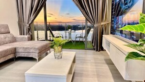 A modern living room with a sofa, a coffee table, and a TV. There is a balcony with a view of the city skyline and a sunset. - Full Sea View Country Garden Danga Bay 11-13pax (Johor Bahru)