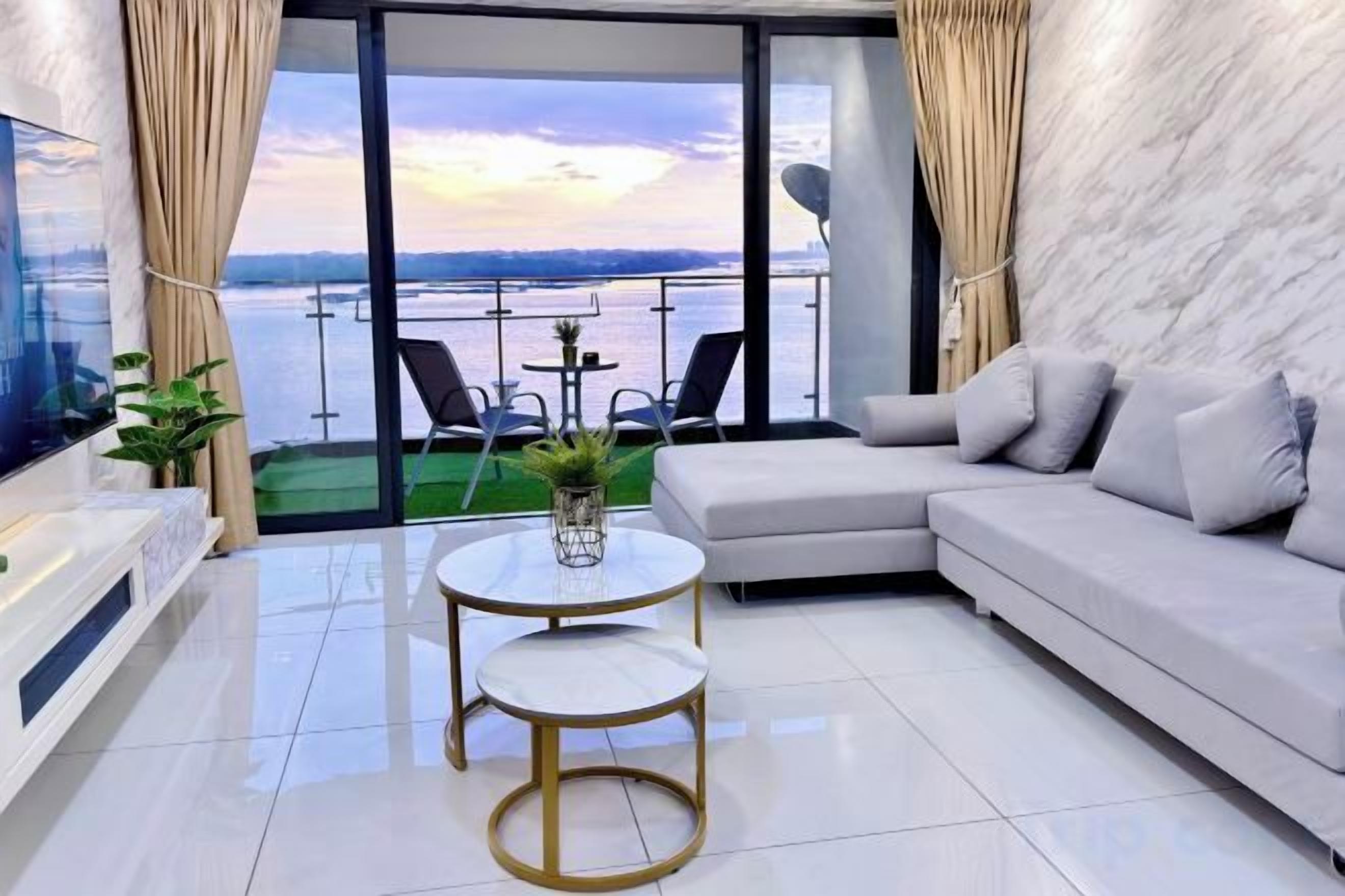 A modern living room with a sofa, a small round table, and a TV. There is a balcony with a view of the water and a bridge.