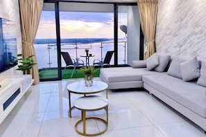 A modern living room with a sofa, a small round table, and a TV. There is a balcony with a view of the water and a bridge. - Full Sea View Country Garden Danga Bay 11-13pax (Johor Bahru)