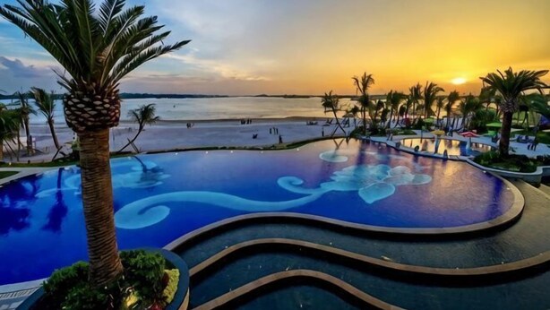 A swimming pool with a wavy design, palm trees, and a view of the beach at sunset. - Full Sea View Country Garden Danga Bay 11-13pax (Johor Bahru)