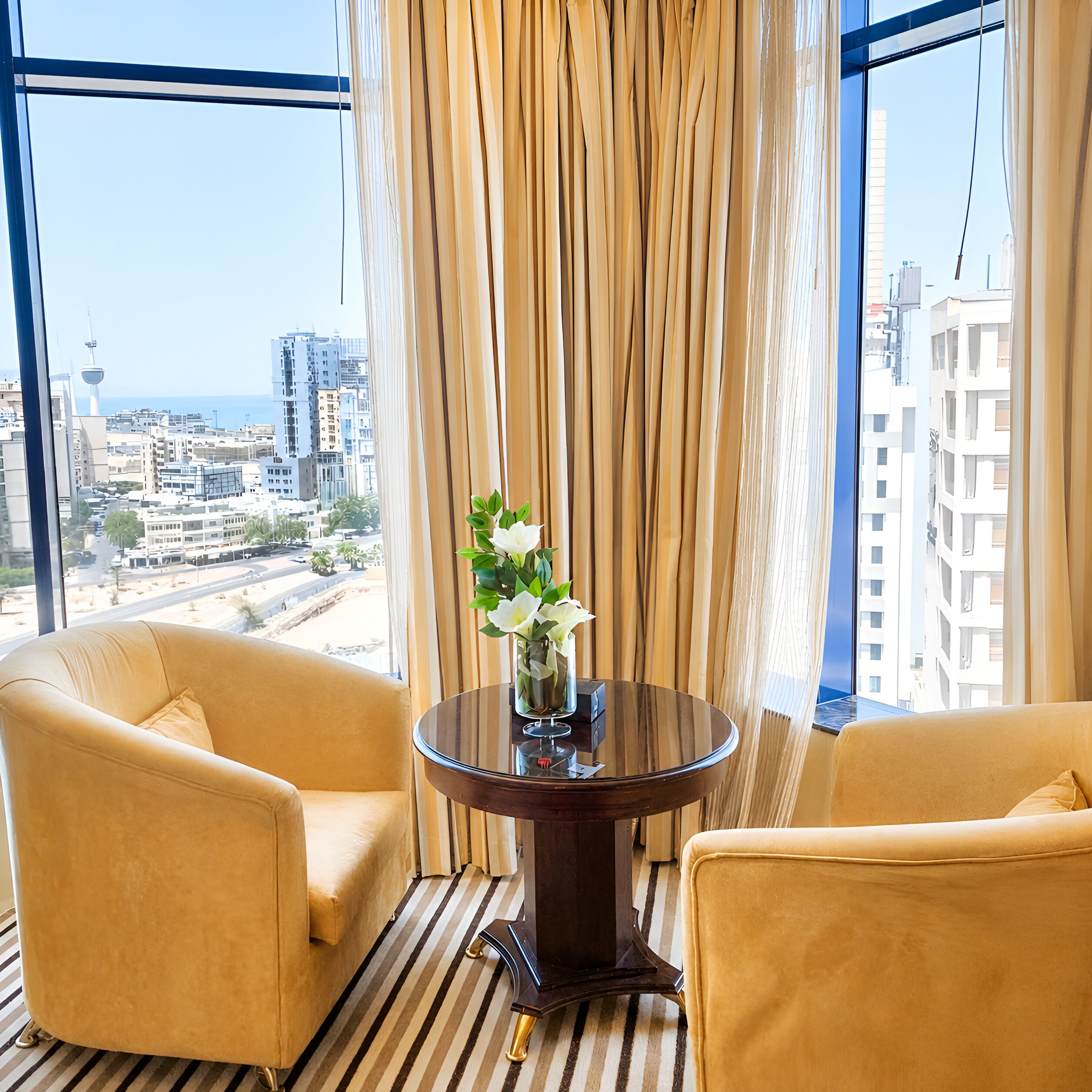 Una habitación con dos sillones amarillos, una mesa redonda con un jarrón de flores blancas y vistas a un paisaje urbano a través de grandes ventanales.