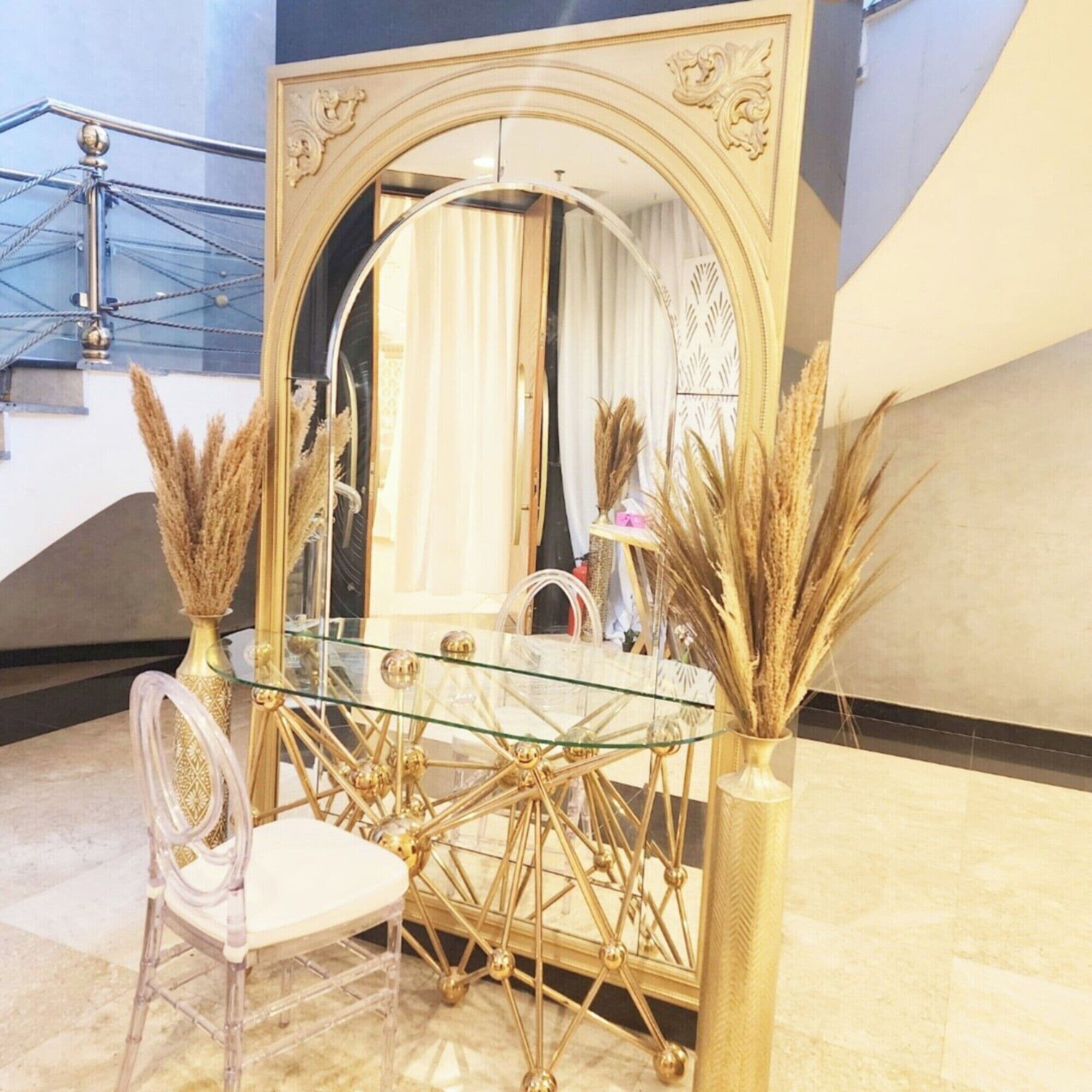 A dining area with a glass table, gold-framed mirror, and decorative plants.