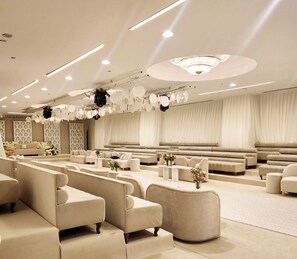 A spacious, well-lit room with multiple seating areas, including sofas and armchairs, arranged around a central area with a large window. - Bloom Hotel (Kuwait City)