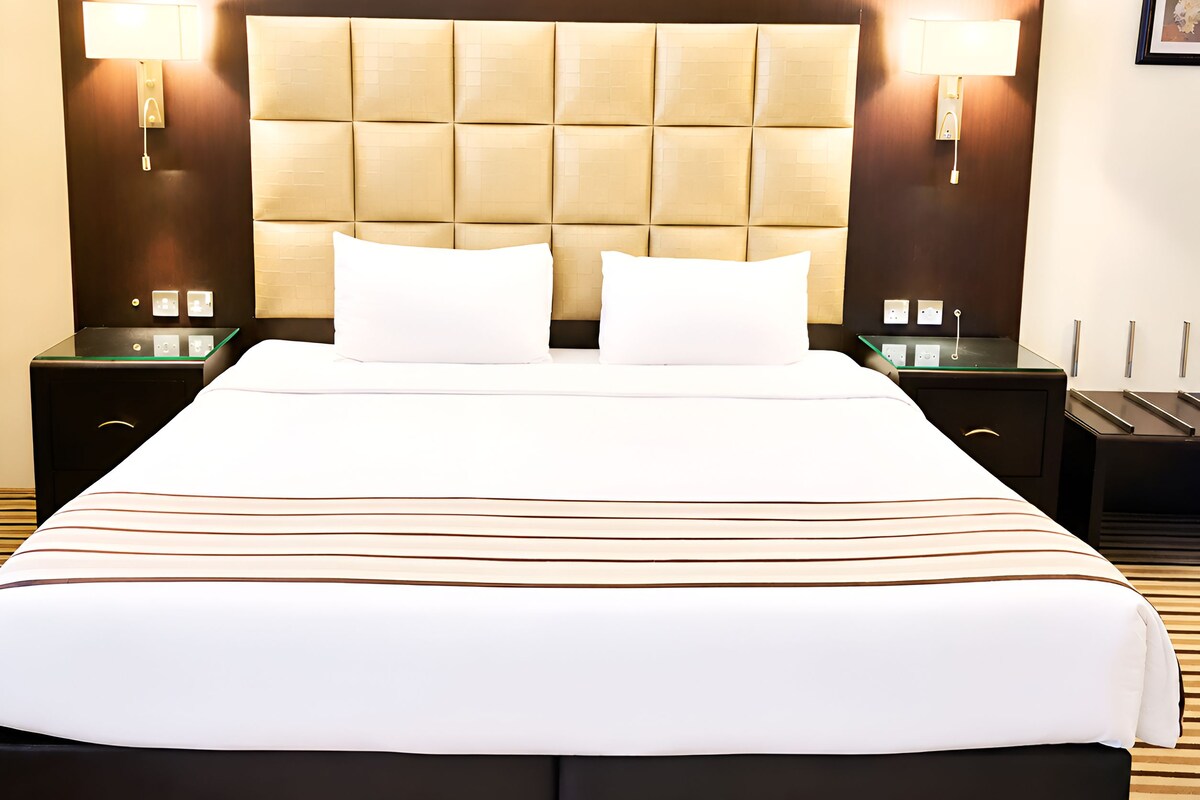 Una habitación de hotel con una cama grande, dos mesitas de noche, un cabecero y lámparas fijadas a la pared.